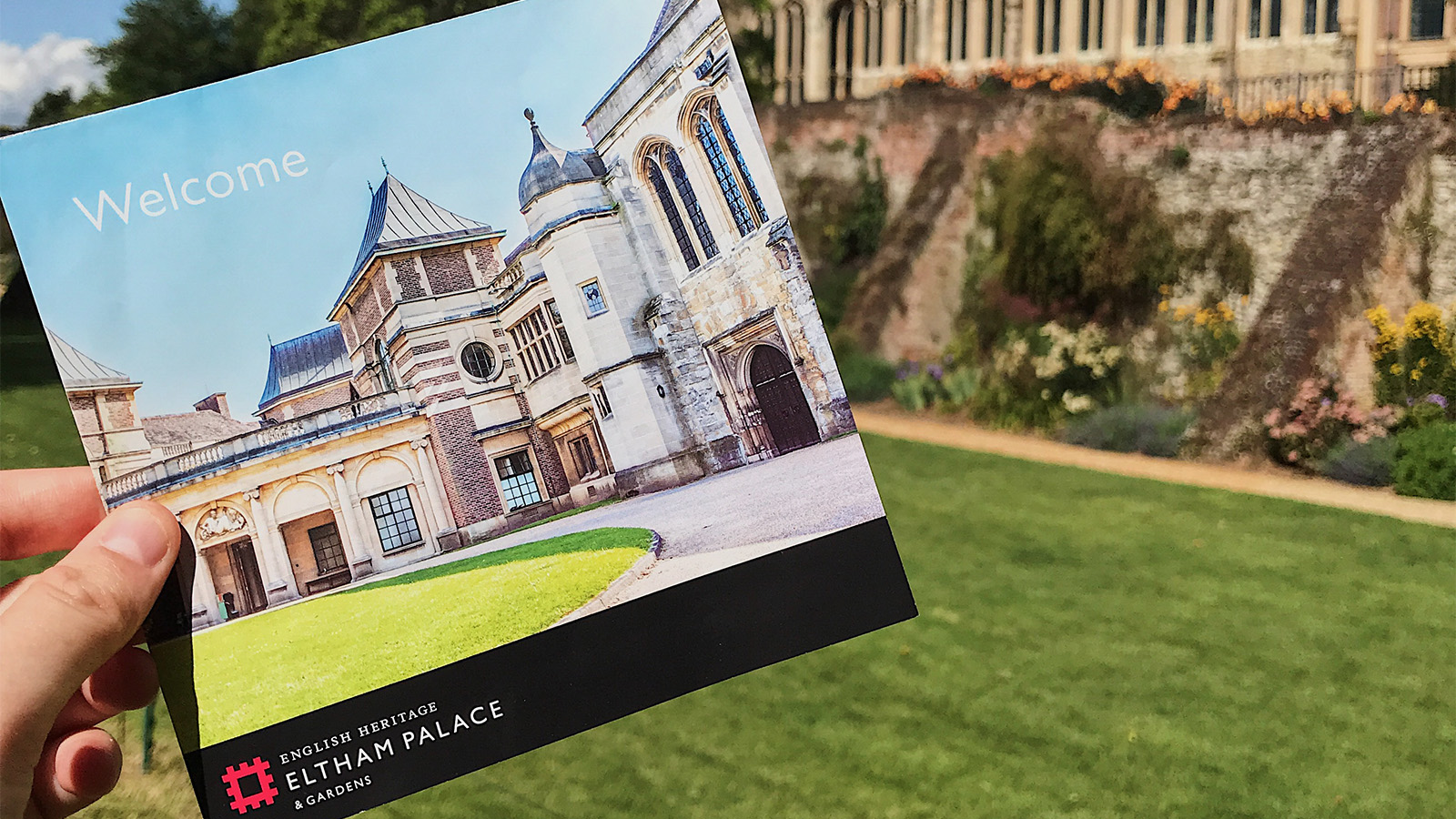 Eltham Palace leaflet held up in front of Eltham Palace on a sunny day